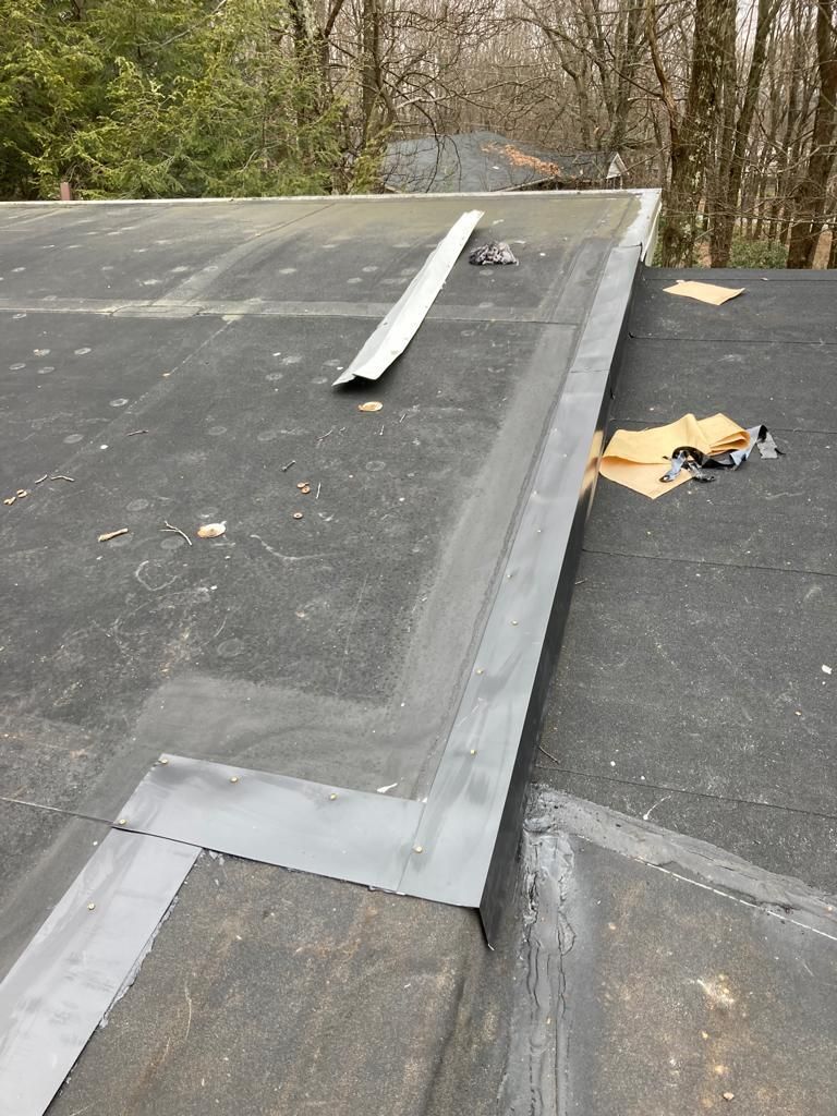 Flat black roof with a loose white metal flashing strip near a seam, outdoors by trees.