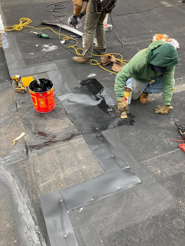 Workers repairing a flat rooftop, applying dark sealant near a red bucket and tools.