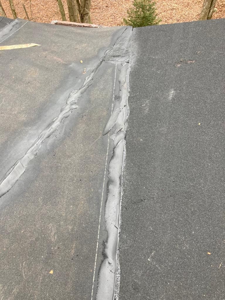 Close-up of a cracked asphalt roof seam with a gray patch and pine trees in the background
