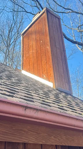 Wooden chimney on a shingled roof against a clear blue sky and bare tree branches
