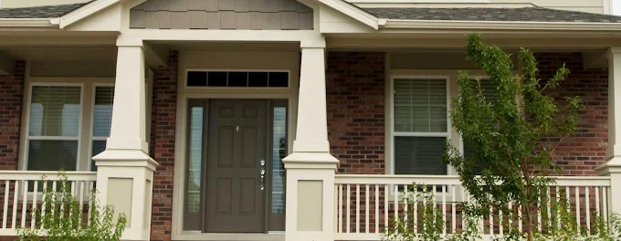 Front porch of a brick house with white columns, railings, and a dark front door.