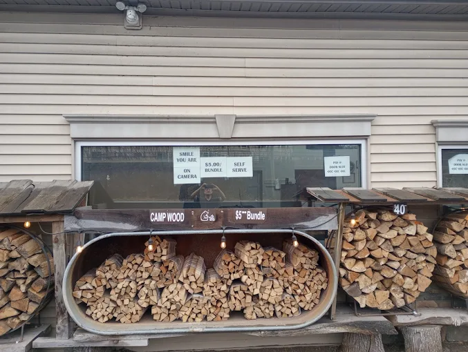 Woodpile stacked beneath a house window, with split firewood stored in open bins and under a metal cover.