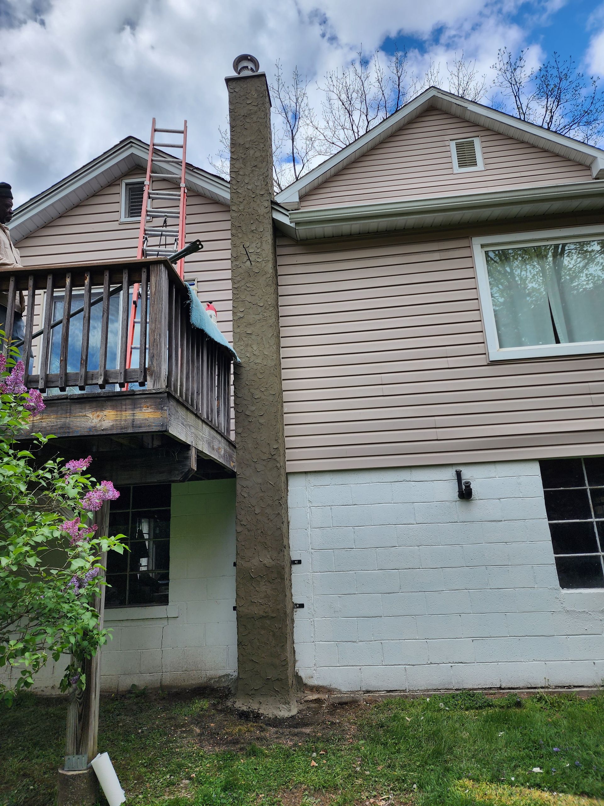 Two-story house exterior with a wooden balcony, central chimney, and a large tree in front.