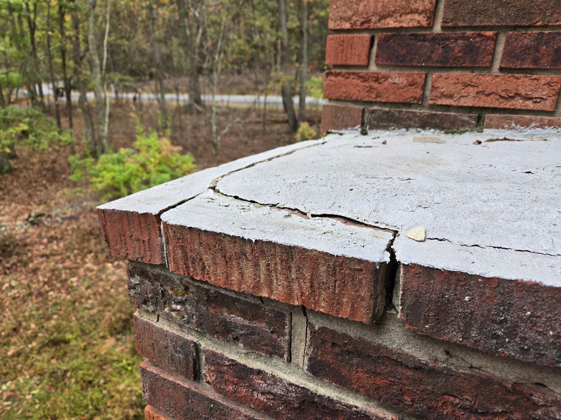 Cracked white concrete porch ledge on a brick house, with wooded yard in the background