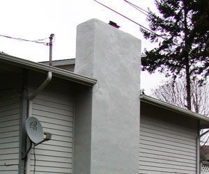 White chimney on a gray house roof with a satellite dish and bare trees in the background