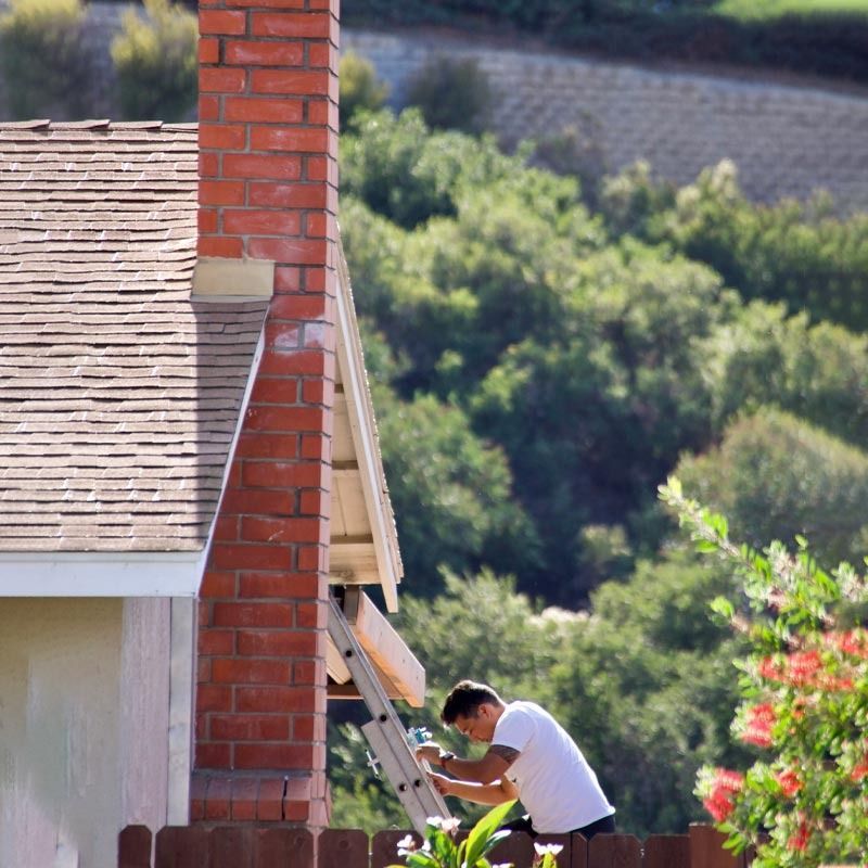 Person painting a house exterior beside a red brick chimney, with green hills in the background
