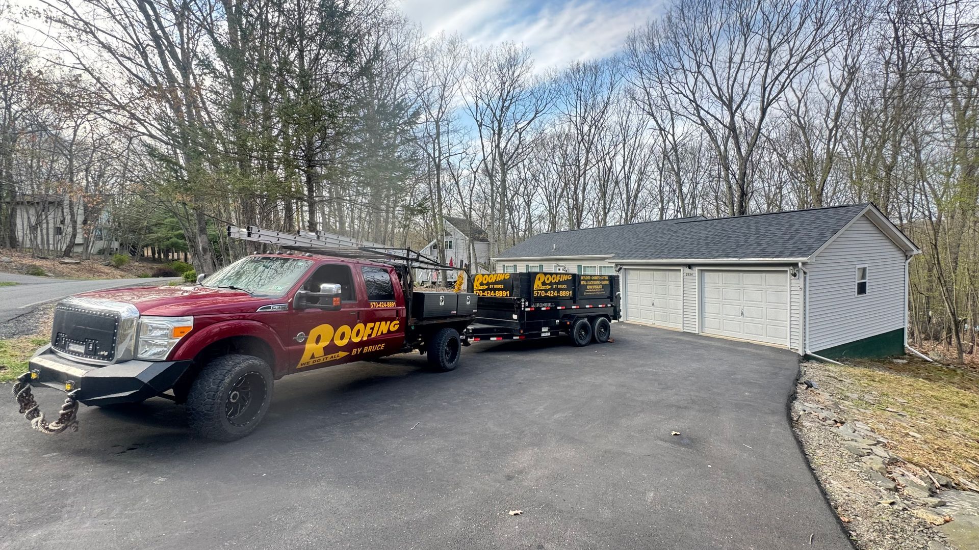 Red tow truck with trailer parked beside a white garage on a driveway, wooded yard in the background