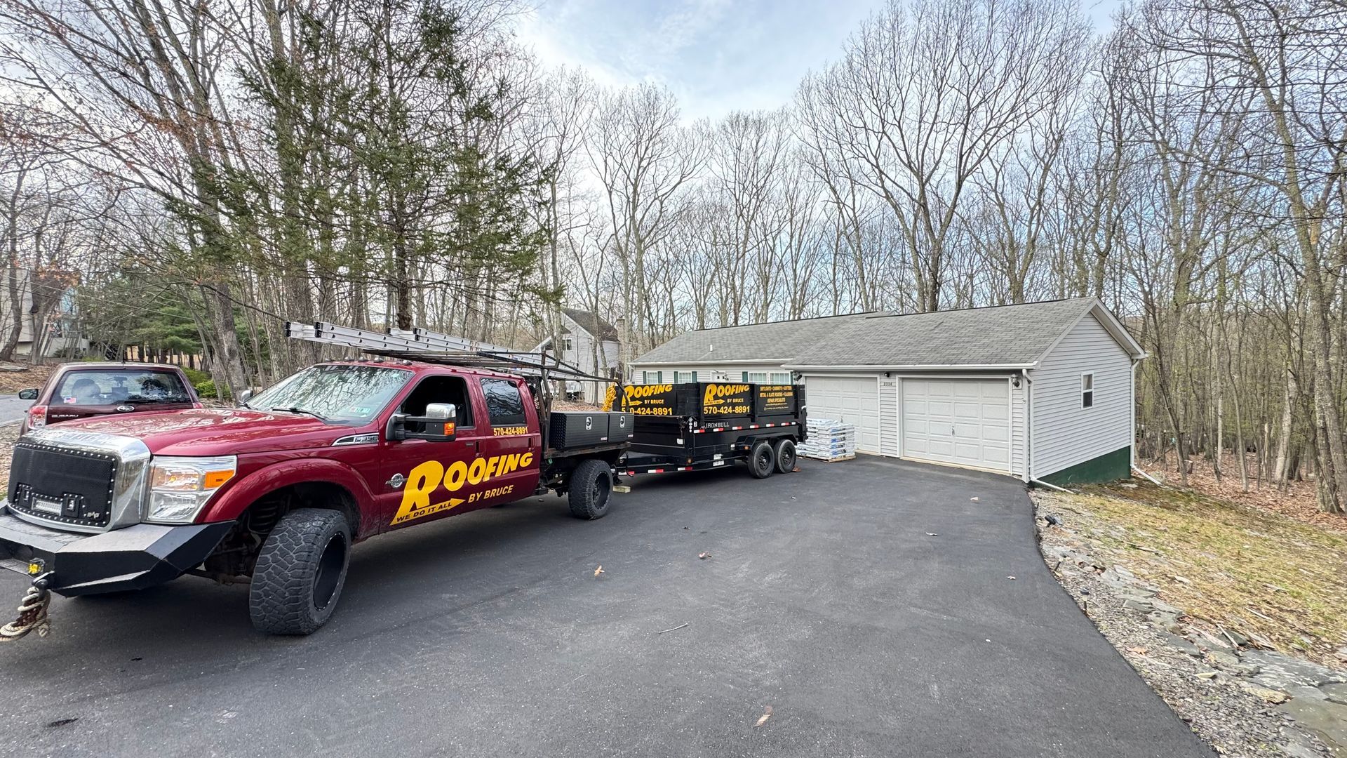 Red tow truck with trailer parked on a driveway beside a white garage in a wooded area