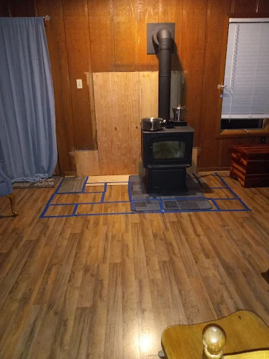 Wood-paneled room with a black wood stove centered on a blue mat and curtains on the left.
