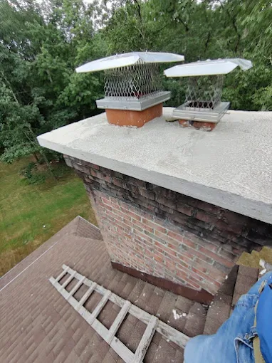 Brick chimney on a rooftop with two metal chimney caps and a ladder leaning against it.