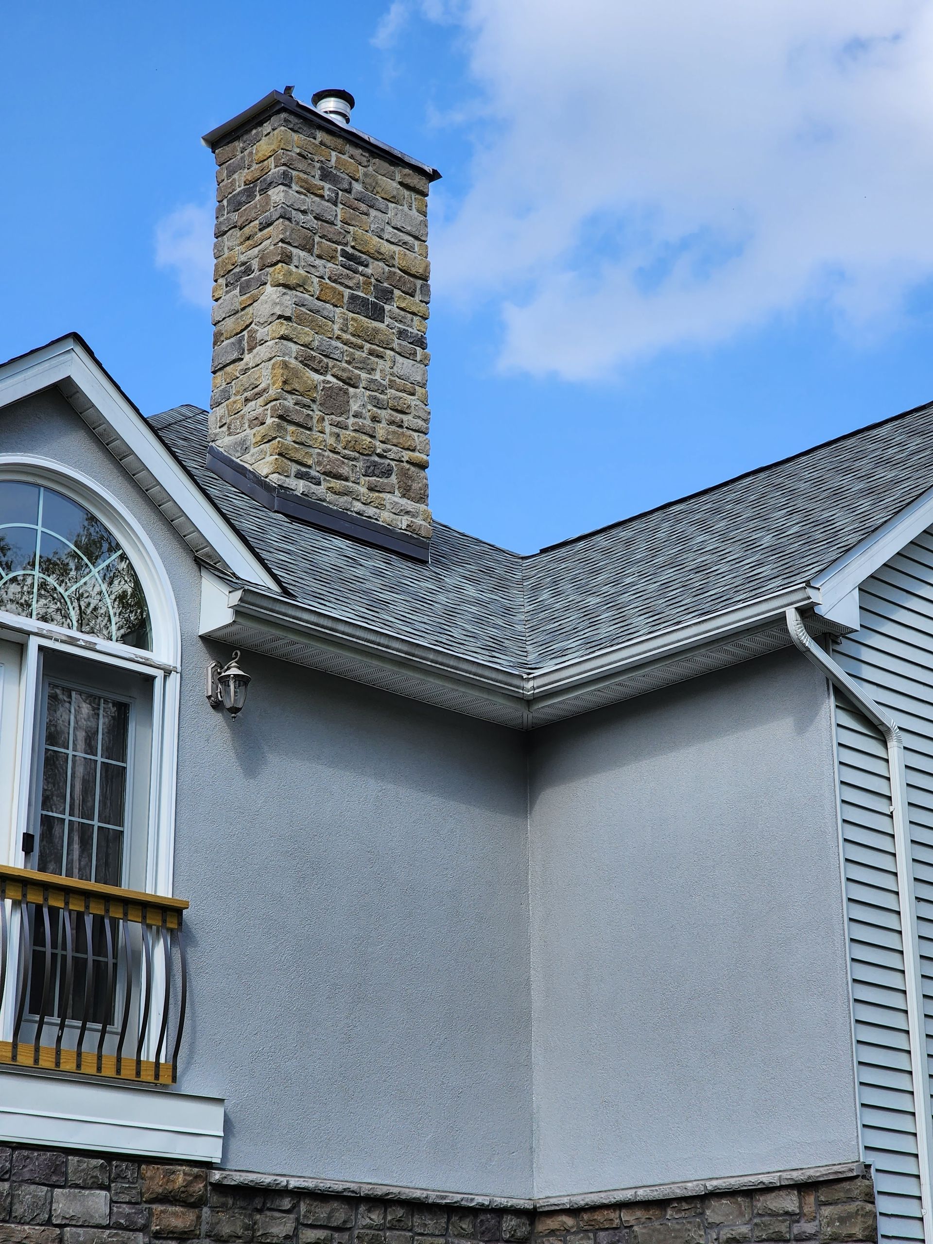 Exterior corner of a gray house with a stone chimney and dormer window against a blue sky