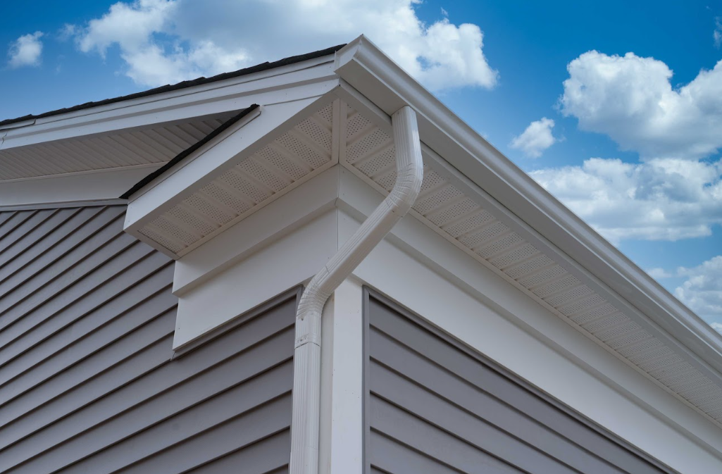 White house corner with vinyl siding, roof eaves, and rain gutter against a blue sky