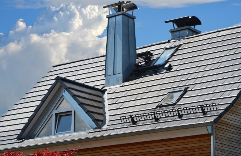 Modern house roof with gray shingles, skylights, and two chimneys under a blue sky