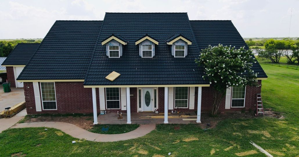 Roofer's trailer and crew near a house with a partially exposed roof and supplies on the ground, sunny day.