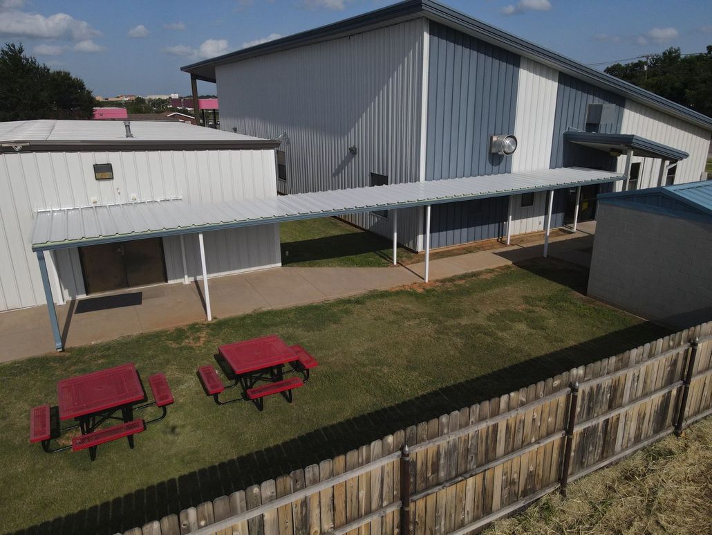 A white and blue metal building with a covered walkway, picnic tables, and a wooden fence.
