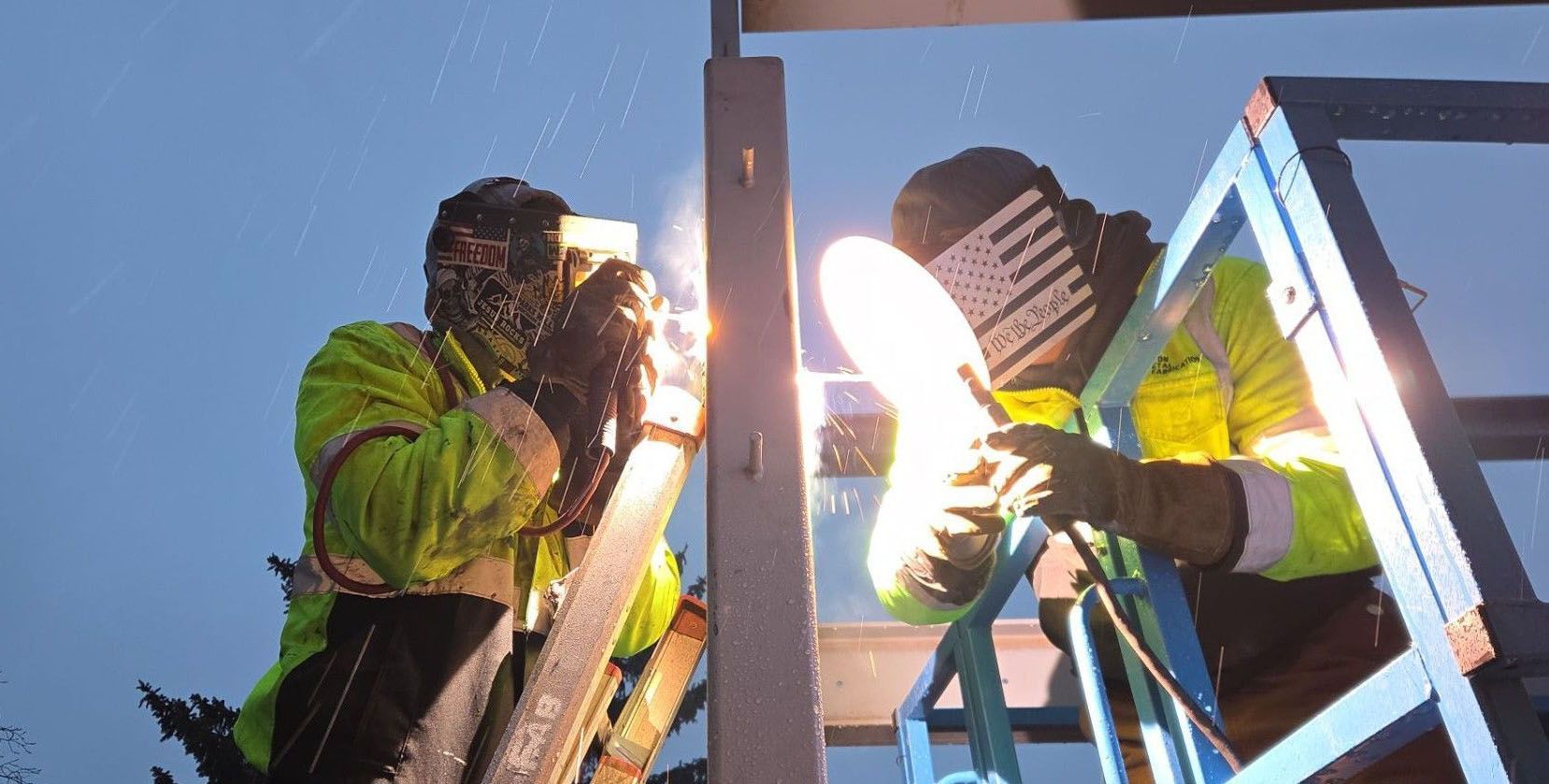 Two workers in high-visibility jackets use welding torches on a metal frame, creating bright light sparks at dusk.