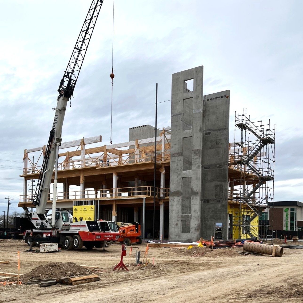 A crane at a construction site with wooden framework, concrete wall sections, and scaffolding under a cloudy sky.