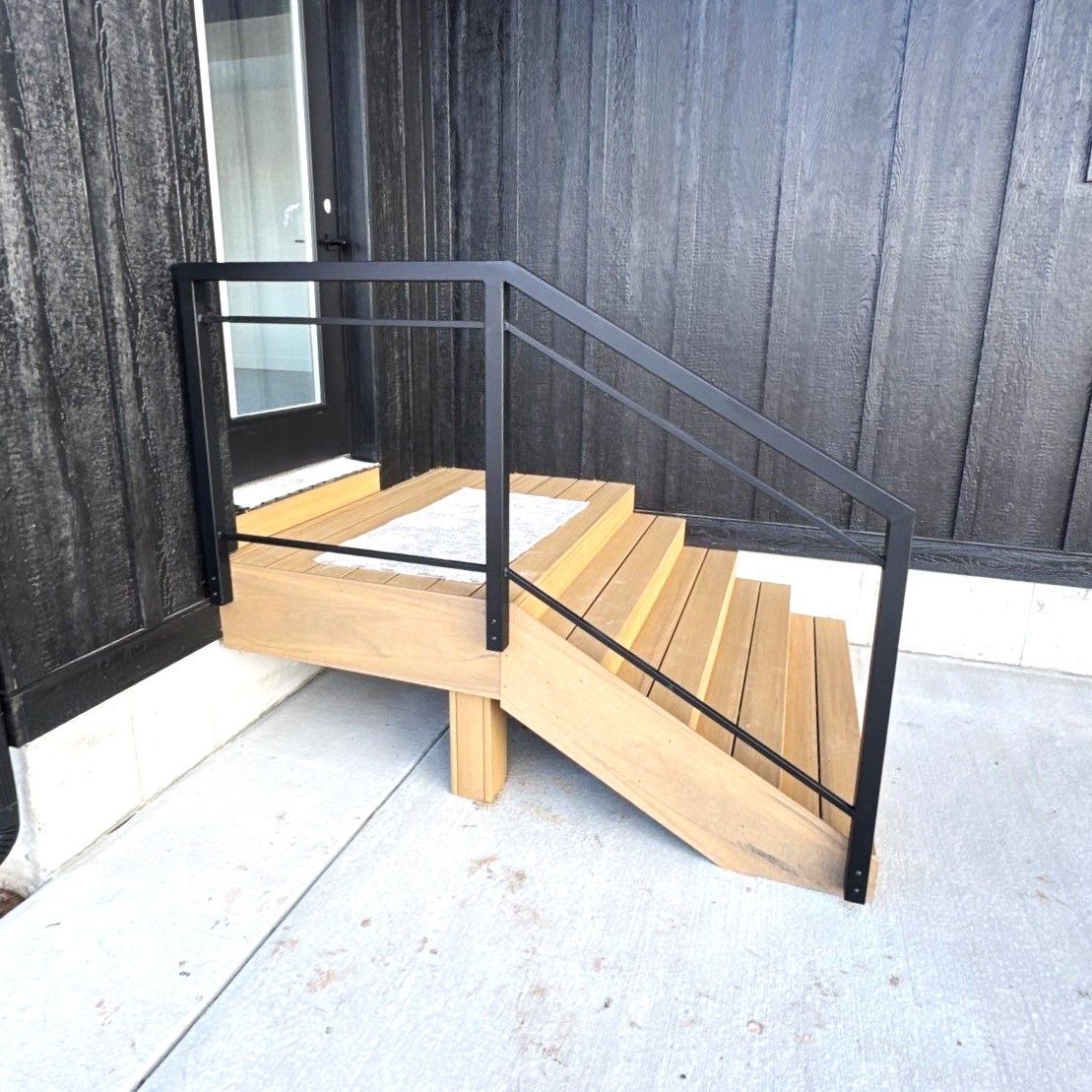 Wooden stairs with a black metal handrail leading to a doorway on a building with black vertical siding.