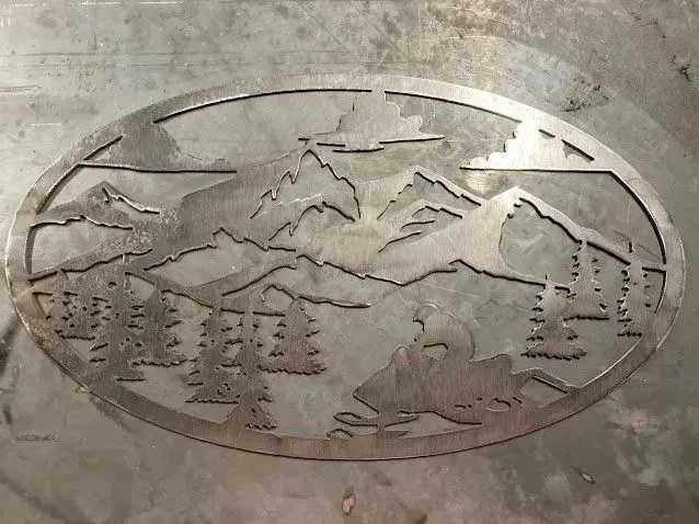 A metal cut-out plaque in an oval frame featuring a snowmobiler riding through a forest with mountains in the background.