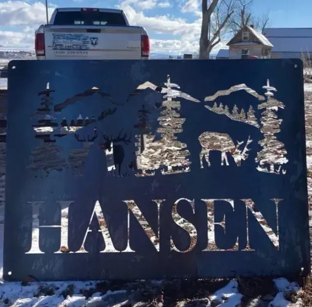 A metal sign with a silhouette of trees, mountains, and a deer above the name 