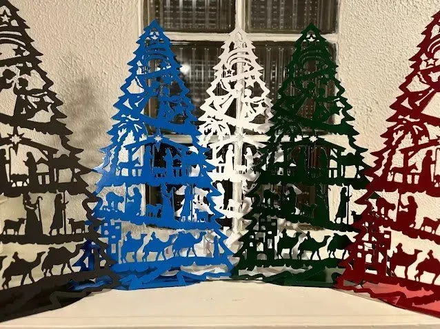 Five decorative Christmas tree cutouts featuring nativity scenes, standing in black, blue, white, green, and red.
