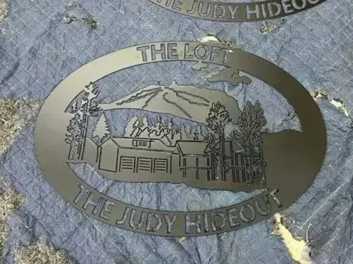 A dark, oval-shaped metal sign with a cut-out illustration of a cabin in front of a mountain, labeled 