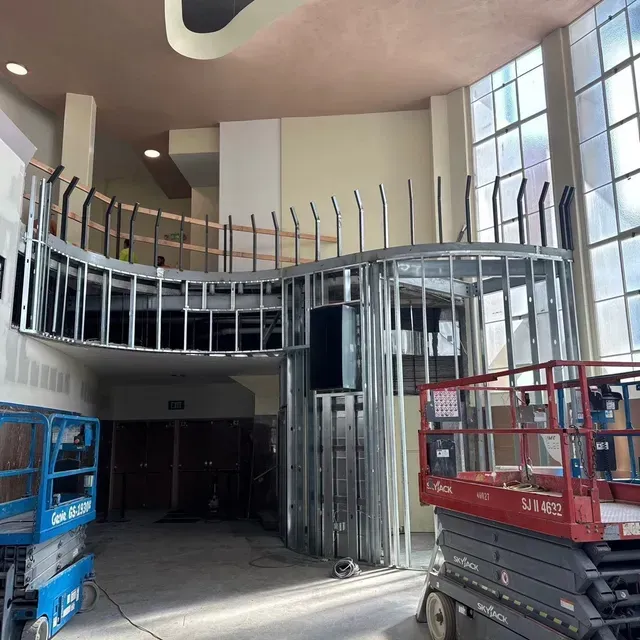 Construction interior with metal framing for a curved balcony, two scissor lifts, and tall windows on the right.
