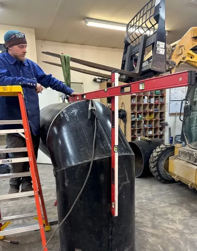 A person uses a T-shaped level tool to check the vertical alignment of a large black metal pipe in a workshop.