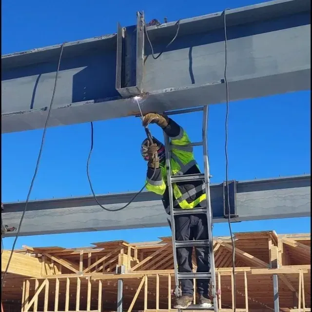 A construction worker in a high-visibility jacket stands on a ladder, using a welding torch on a steel I-beam.
