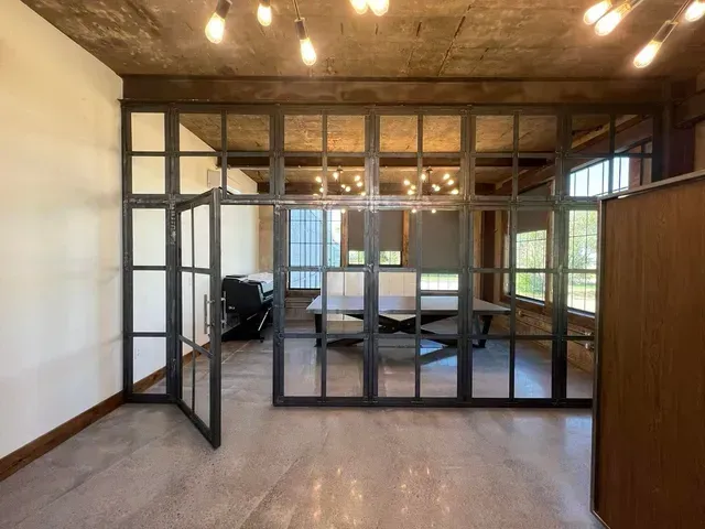 An office space with polished concrete floors, a glass and dark metal partition, and an open door leading to a desk.