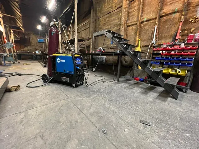 A blue Miller welding machine with a gas cylinder in a workshop near a metal staircase and shelving unit.