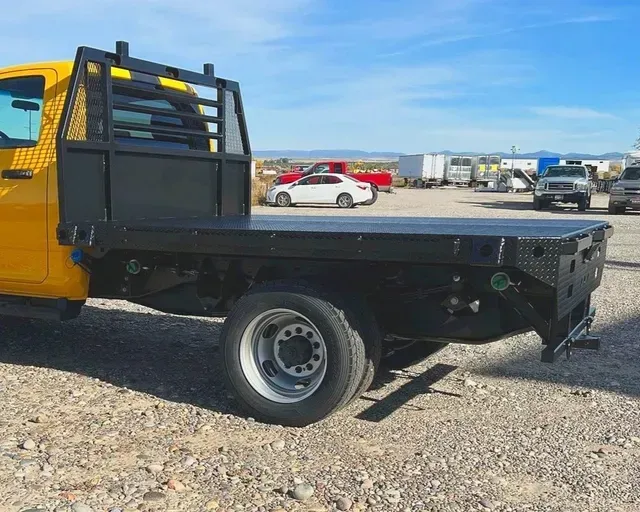 A bright yellow truck with a black flatbed utility body parked in a gravel lot on a sunny day.