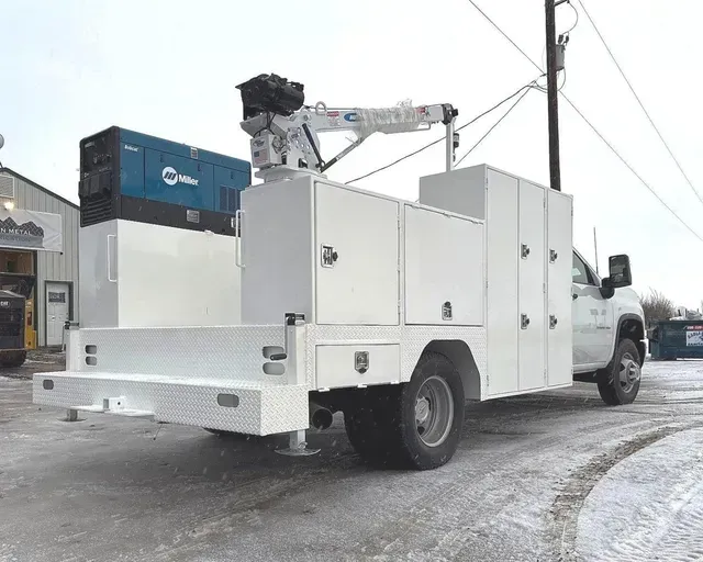 A white utility service truck with a mounted crane and a blue Miller generator parked on a snowy lot.