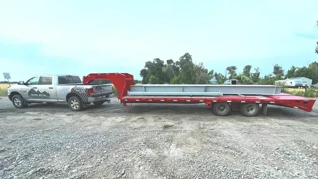 A silver pickup truck towing a long red flatbed trailer carrying a large metal beam on a gravel lot under a cloudy sky.