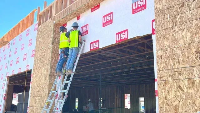 Two workers in high-visibility vests on a ladder installing white weather barrier wrap on a wooden building frame.