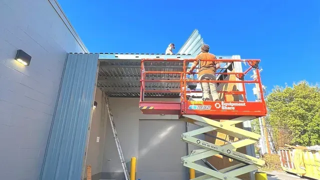 Workers install corrugated metal roofing on a building exterior using a red scissor lift and a ladder under a blue sky.
