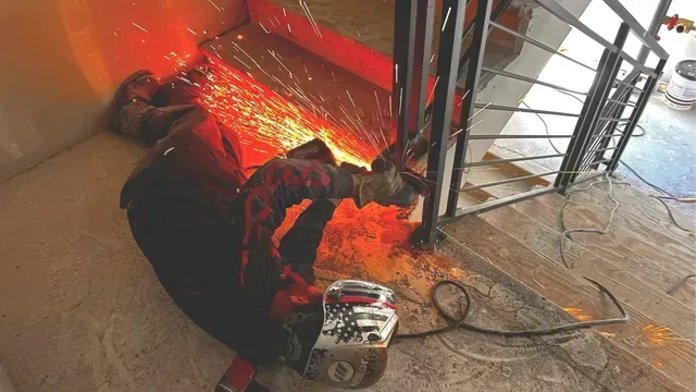 A worker wearing a welding helmet uses an angle grinder to cut a metal railing, producing a shower of bright sparks.
