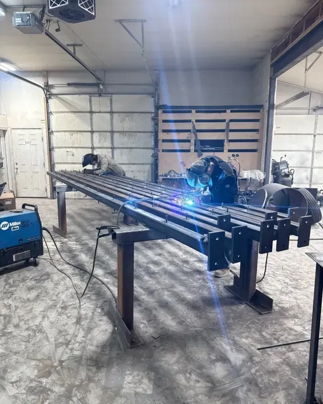 Two welders work on a long metal structure in a workshop, sparks flying as one joins steel components on a workbench.