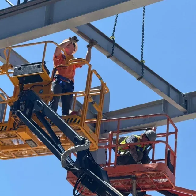Two construction workers in high-visibility gear stand in aerial work platforms, installing steel beams against a blue sky.