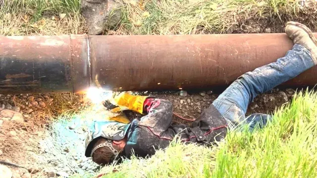 A welder in protective gear lies in a trench, welding a large metal pipeline outdoors.