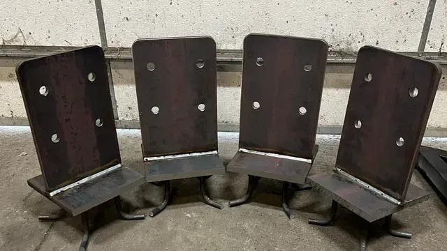 Four steel brackets with flat vertical plates and bolted holes arranged in a semi-circle on a concrete floor.
