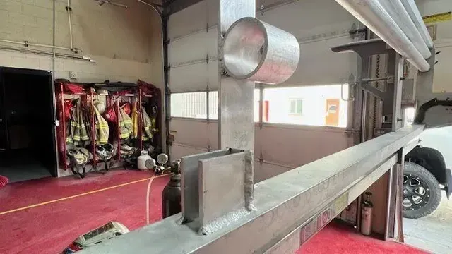 Inside a fire station, a metal beam with a mounted bracket stands near gear racks and a vehicle parked by a garage door.