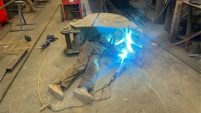 A person in work clothes lies on a shop floor, welding the metal interior of a large, heavy-duty excavator bucket.