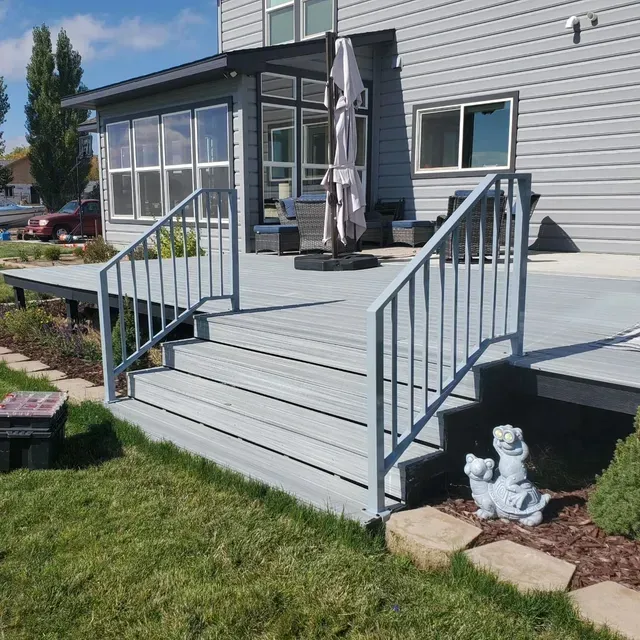 A light gray backyard deck with stairs, a railing, and a patio area outside a house with gray siding.