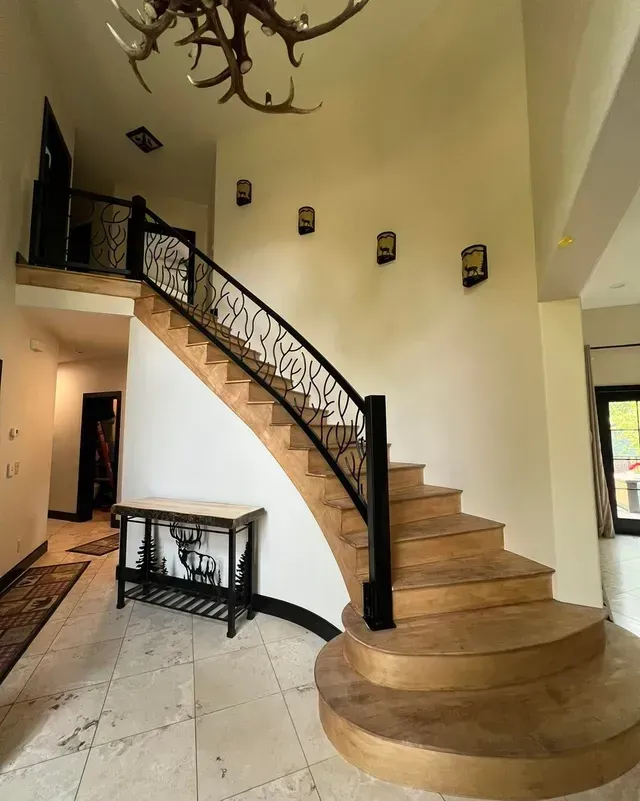 An elegant curved staircase with wood steps and iron railings in a grand foyer with a large antler chandelier.