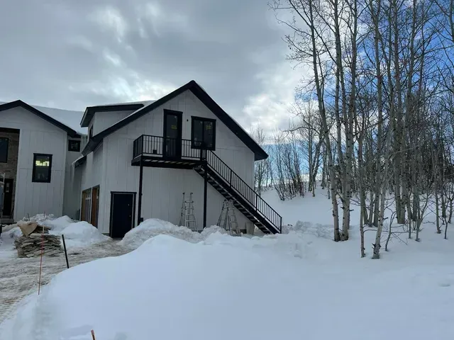 A modern white house with black trim and an exterior metal staircase sits in a snow-covered landscape with bare trees.