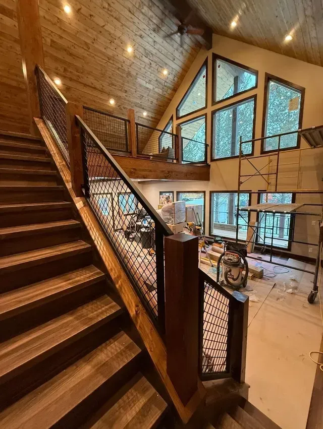 Wooden staircase with metal mesh railing leads to a loft overlooking a living area under construction with large windows.