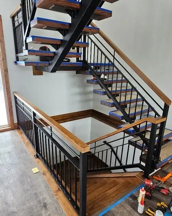 A two-story staircase featuring black metal railings, wooden handrails, and wood treads, partially under construction.