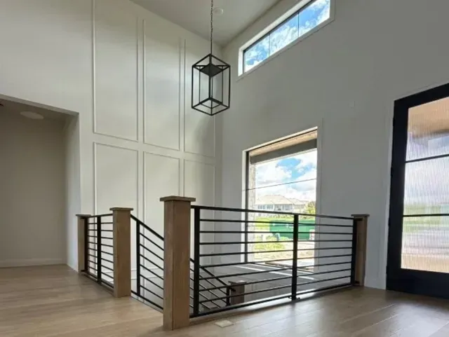 A modern foyer with wood flooring, a black metal stair railing, a tall pendant light, and paneled walls under a window.