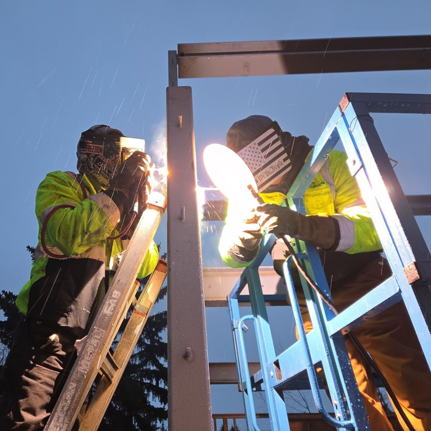 Two construction workers in neon safety vests and welding helmets weld a metal frame outdoors against a blue sky.